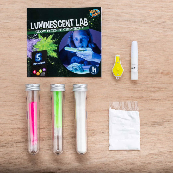 A Luminescent Lab Glow Science Chemistry kit, featuring three test tubes, a powder packet, a small dropper bottle, a yellow clip, and an instruction booklet on a wooden surface—perfect for glowing experiments that reveal the marvels of photoluminescence.